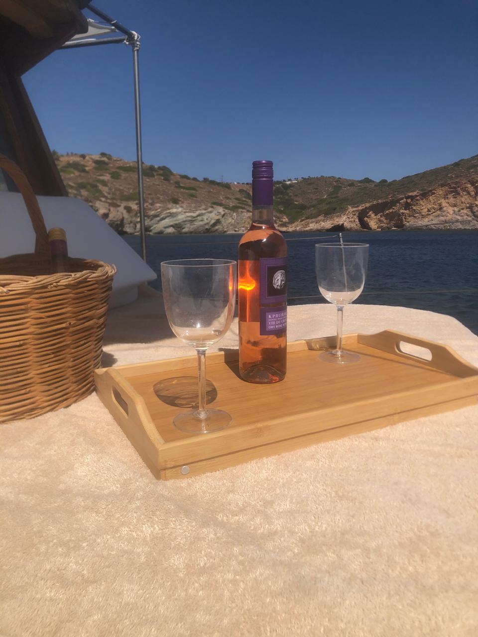 A chilled bottle of rosé and two glasses on a wooden tray, anchored beside a rocky cove